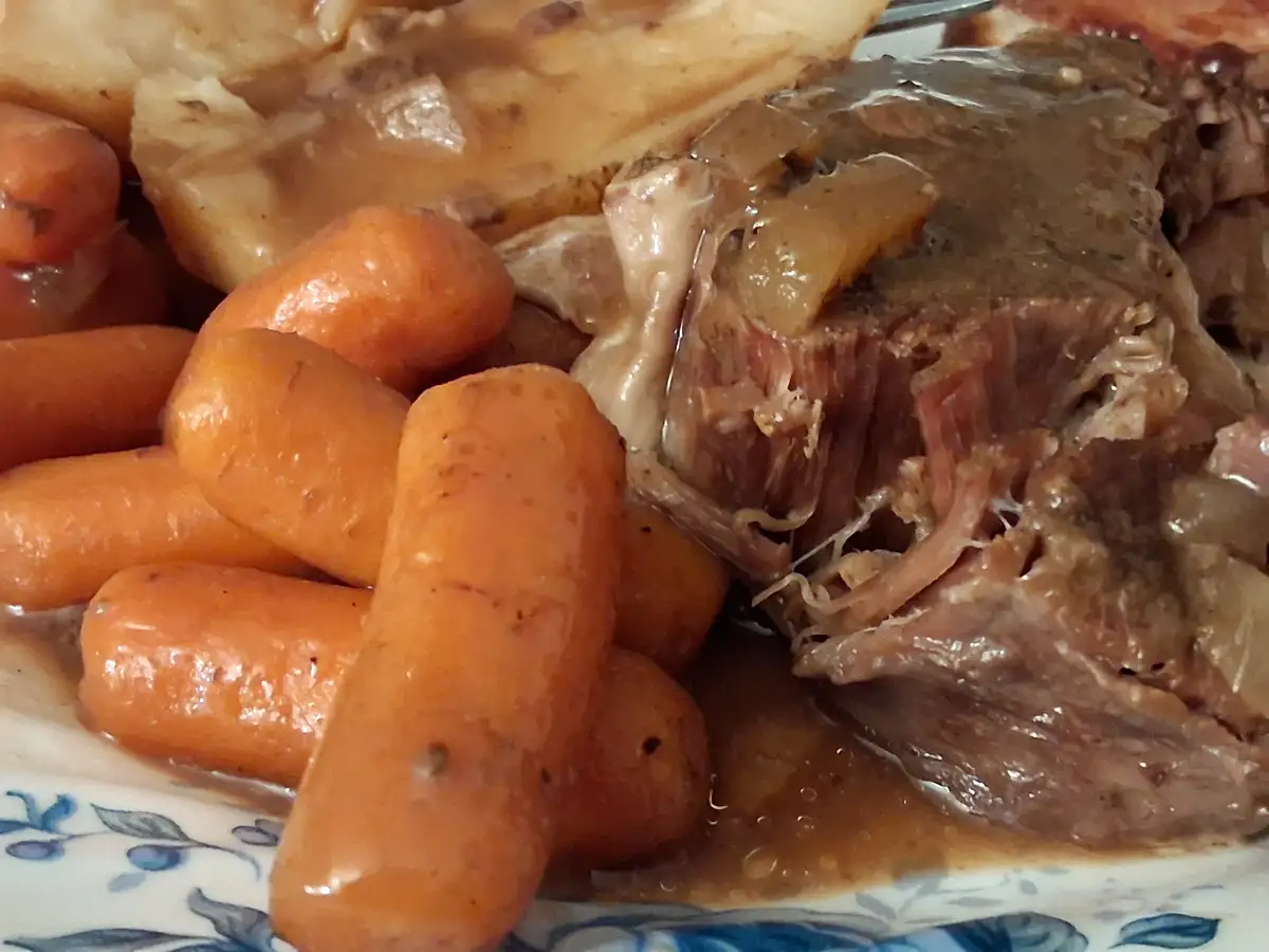 A close-up photo of a plate of food featuring tender, shredded beef covered in gravy, alongside bright orange baby carrots…