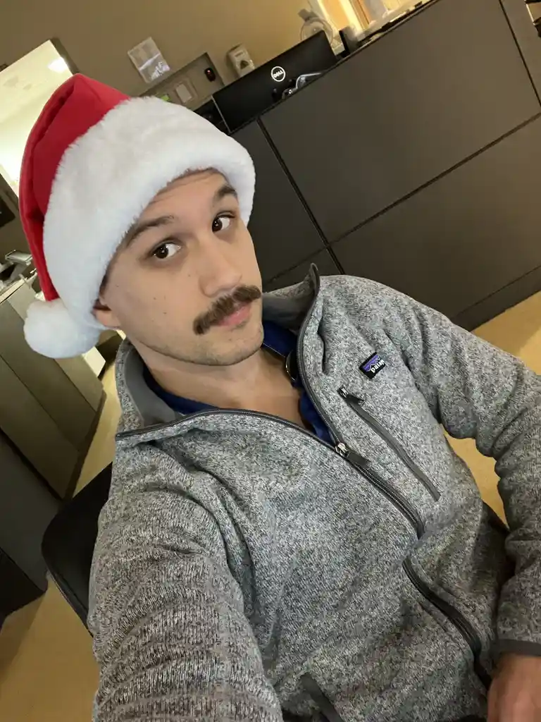 A man wearing a red and white Santa hat and a grey zip-up jacket looks directly at the camera with a slightly confused or…