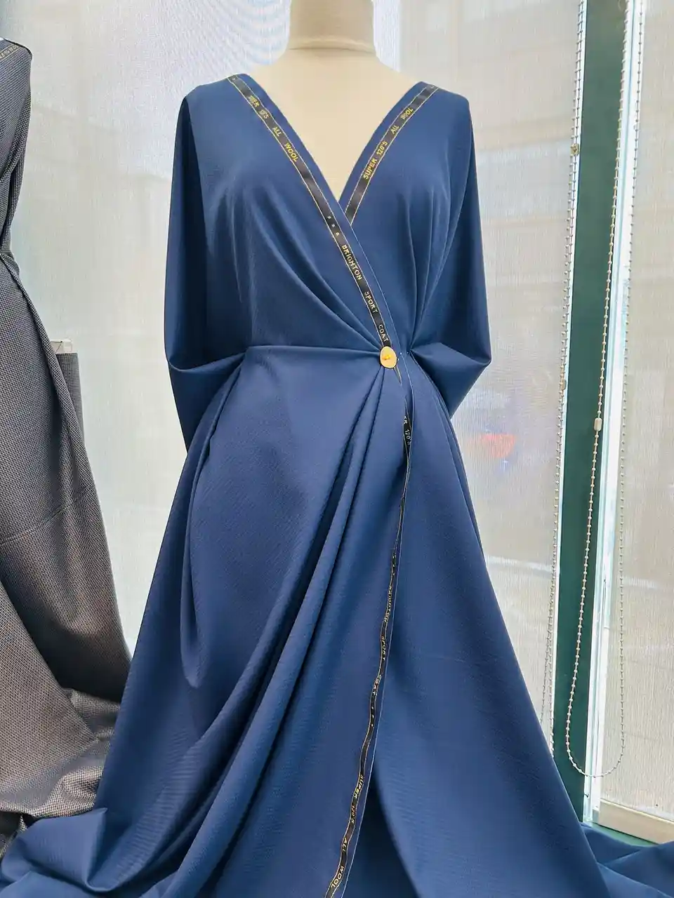 A blue dress on a mannequin features a black ribbon with gold text reading 'super 120s all wool' and 'brighton sport coat'…