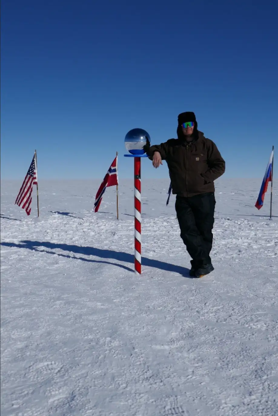 A person in winter gear stands in a snowy, flat landscape next to a red-and-white striped pole topped with a reflective…