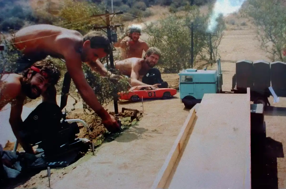 A behind-the-scenes photo from the filming of 'The Dukes of Hazzard' shows shirtless men working on a miniature set with a…