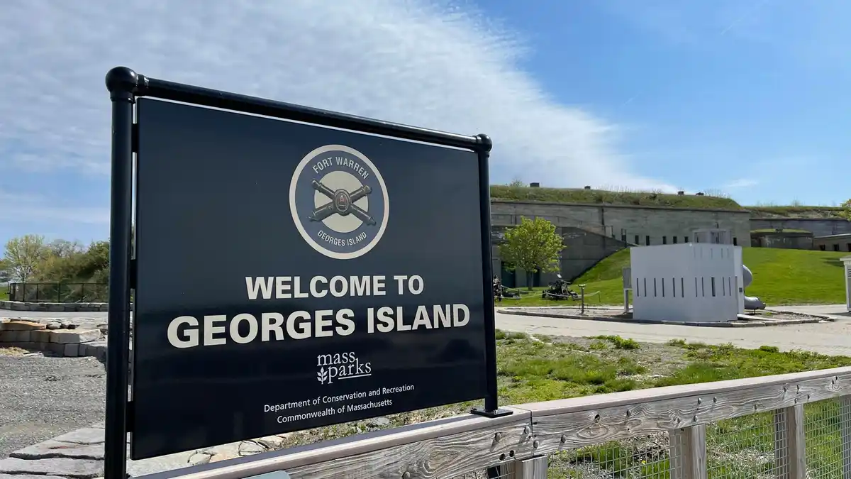 A black welcome sign for Georges Island, managed by MassParks, stands in front of Fort Warren's stone fortifications under a…
