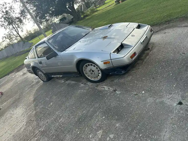 A silver, visibly aged and rusty Nissan 300ZX Turbo is parked crookedly on a cracked asphalt driveway, surrounded by grass…