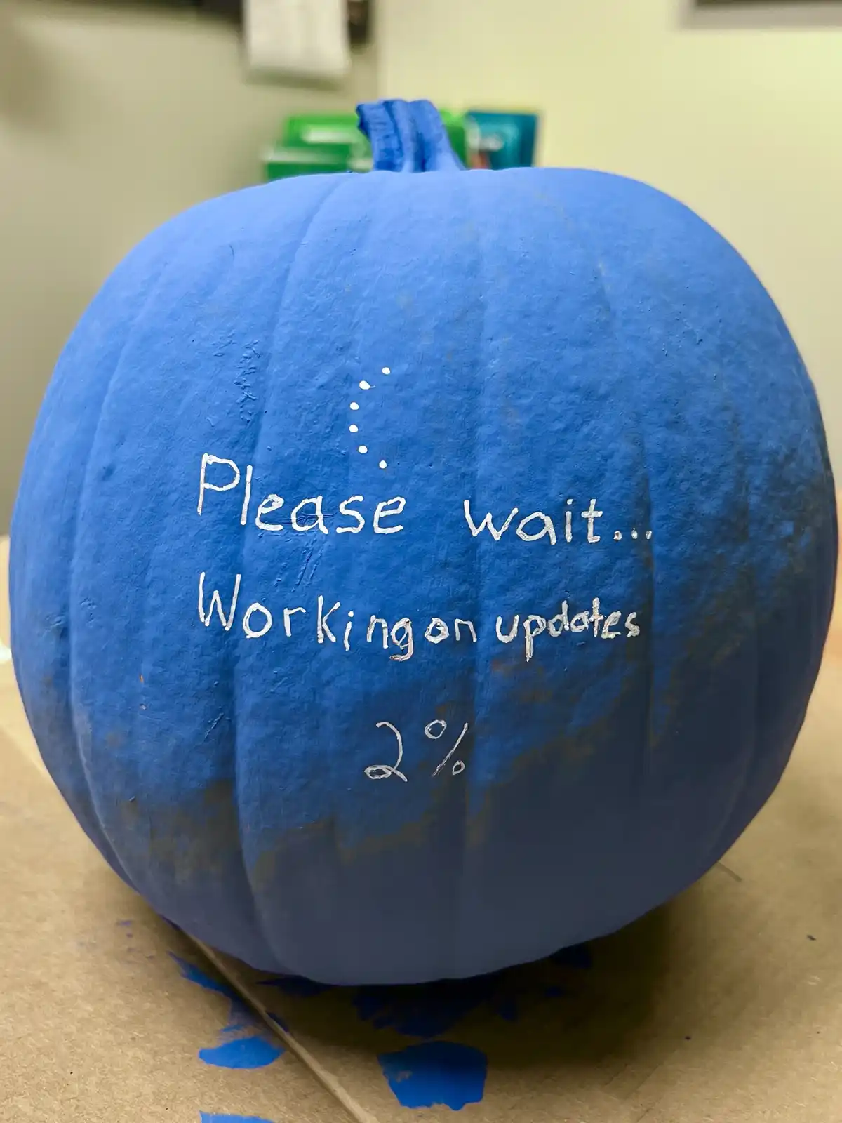 A blue-painted pumpkin with white handwritten text that reads 'Please wait... Working on updates 2%' and a loading dot…