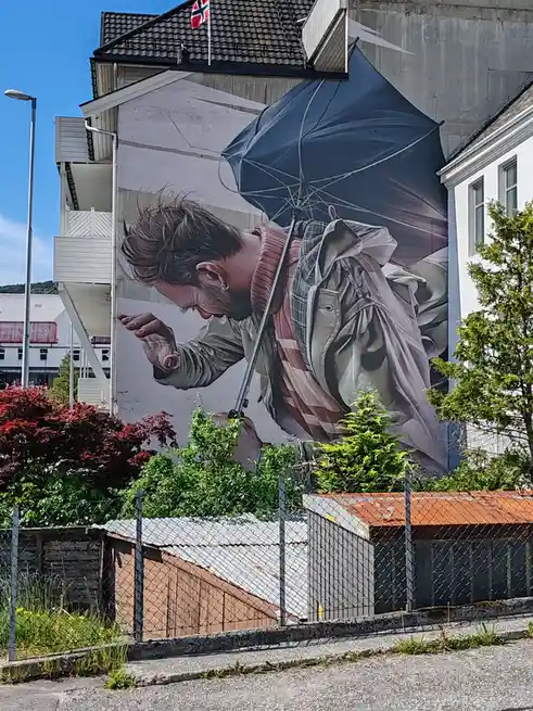 A large, realistic mural on a building wall depicts a man with a beard and ear piercing, holding a dark blue umbrella that…