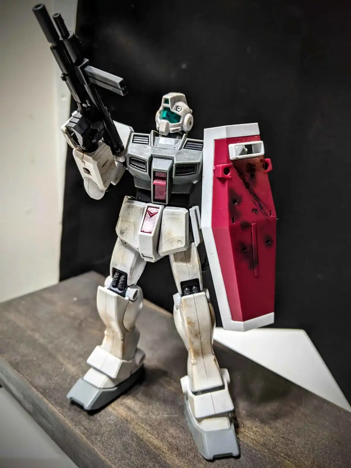 A weathered, white and grey Gundam model stands on a wooden surface holding a black rifle and a red shield with simulated…