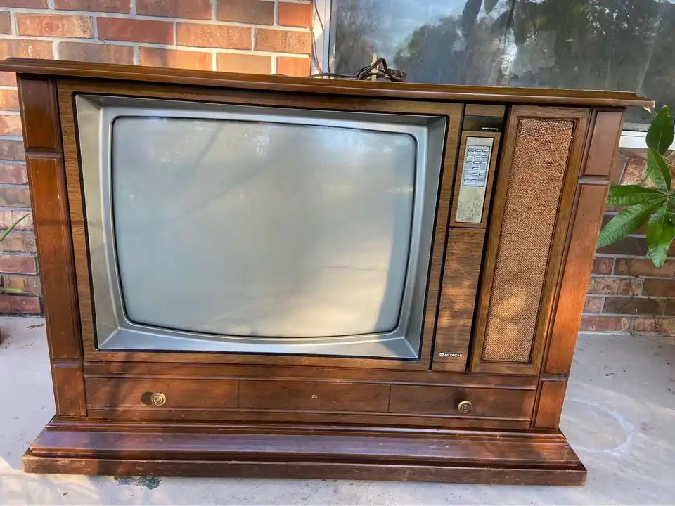 An old wooden Hitachi CRT television with a blank screen sits outdoors on a concrete patio against a brick wall, with the…
