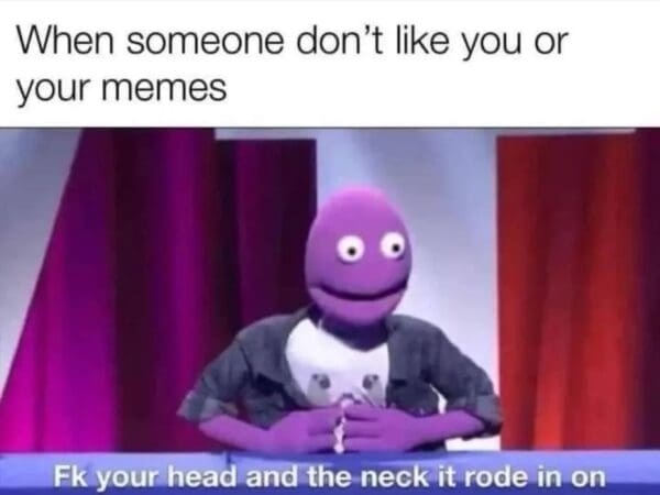 A purple puppet with large white eyes and a wide smile sits at a desk, hands clasped, against a backdrop of red curtains…