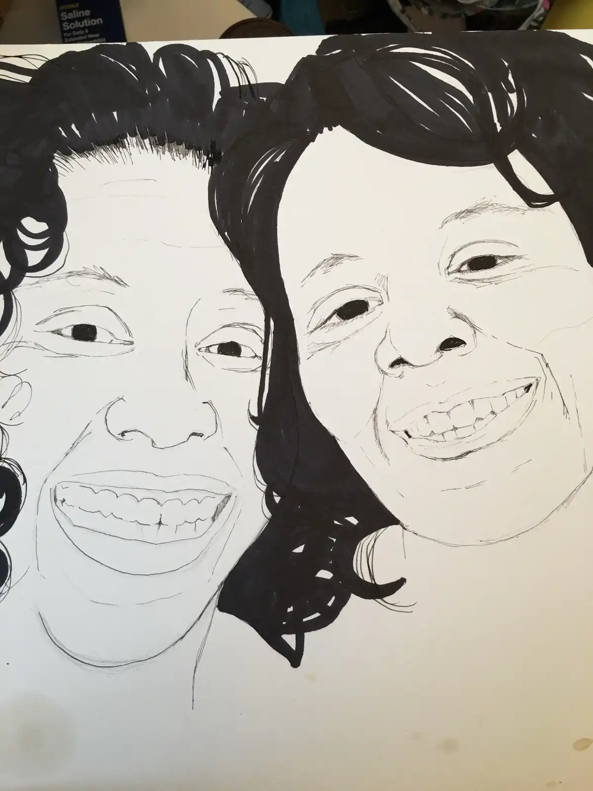 A hand-drawn black-and-white sketch on paper shows two smiling faces side by side, both with dark hair and wide grins. The…