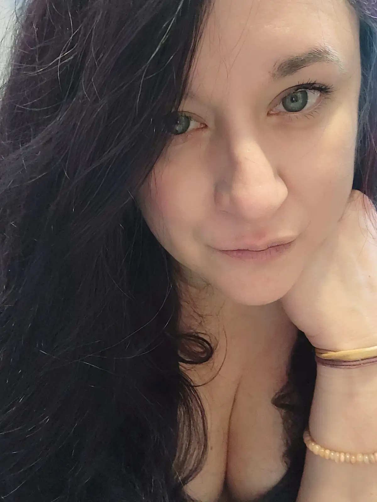 A close-up selfie of a woman with dark hair and green eyes, looking directly at the camera with a subtle smirk while resting…