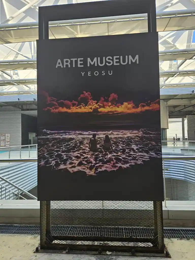 A large black sign for the Arte Museum Yeosu features a scenic photo of two people sitting in ocean waves under a dramatic…