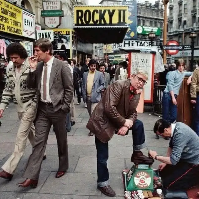 A vintage street scene from the 1970s shows a man in a brown leather jacket sitting on a shoe-shining box while a shoeshine…