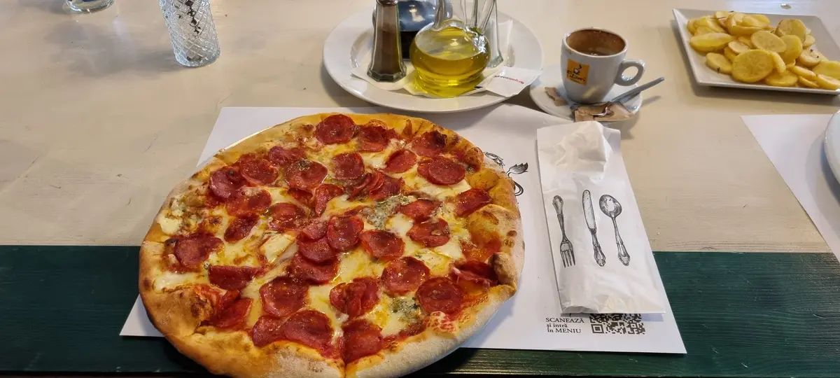 A close-up photo of a pepperoni pizza on a white paper mat on a table, with a cup of coffee, a plate of potato chips, and…