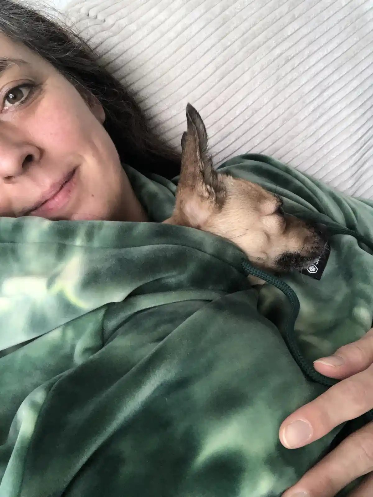 A person with dark hair lies on a light-colored ribbed pillow, wearing a green tie-dye hoodie, with a small tan dog sleeping…