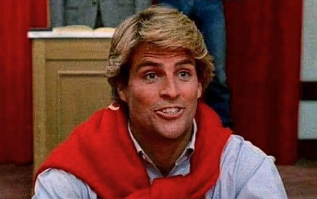A man with blonde hair and a red scarf over a light blue shirt looks directly at the camera with a slightly forced, awkward…