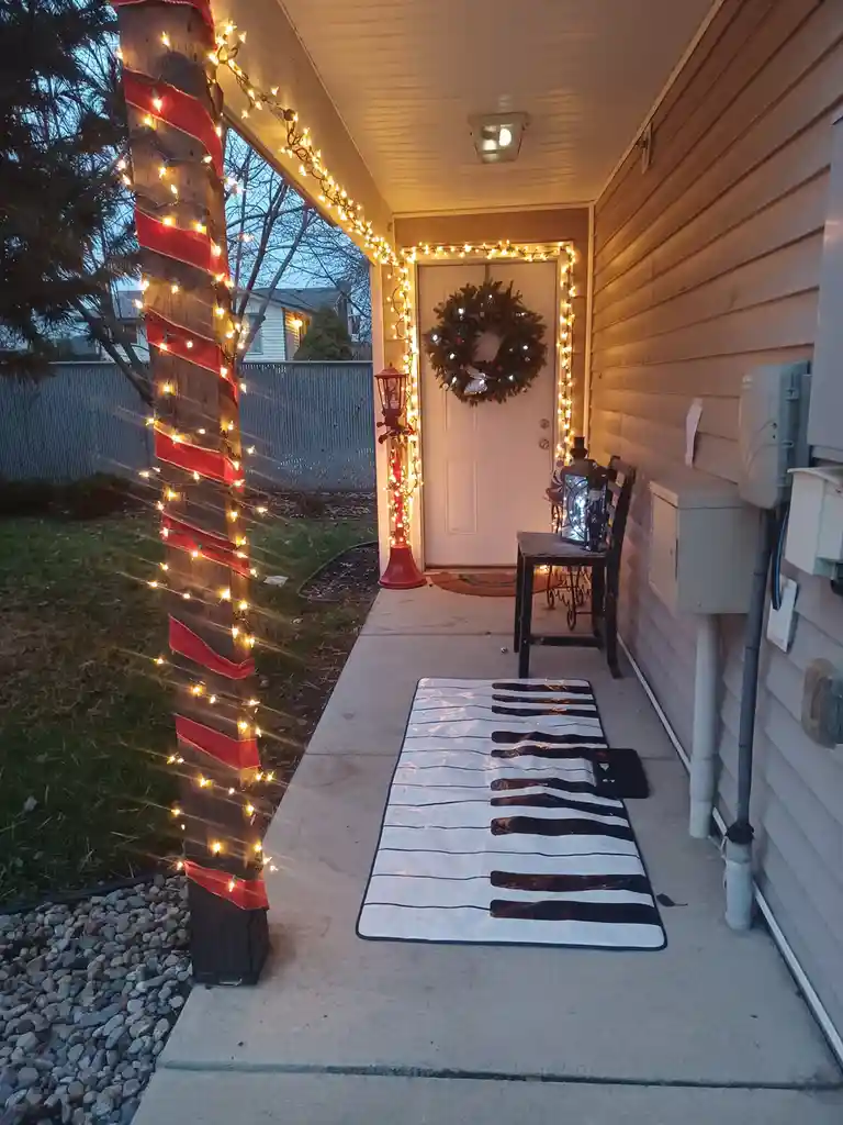 A decorated front porch at dusk features a large piano-key rug laid out on the concrete walkway leading to the door. The…