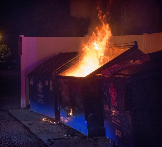 A dumpster is on fire at night, flames shooting out of its open top, with smoke rising. The dumpster is blue and has a logo…