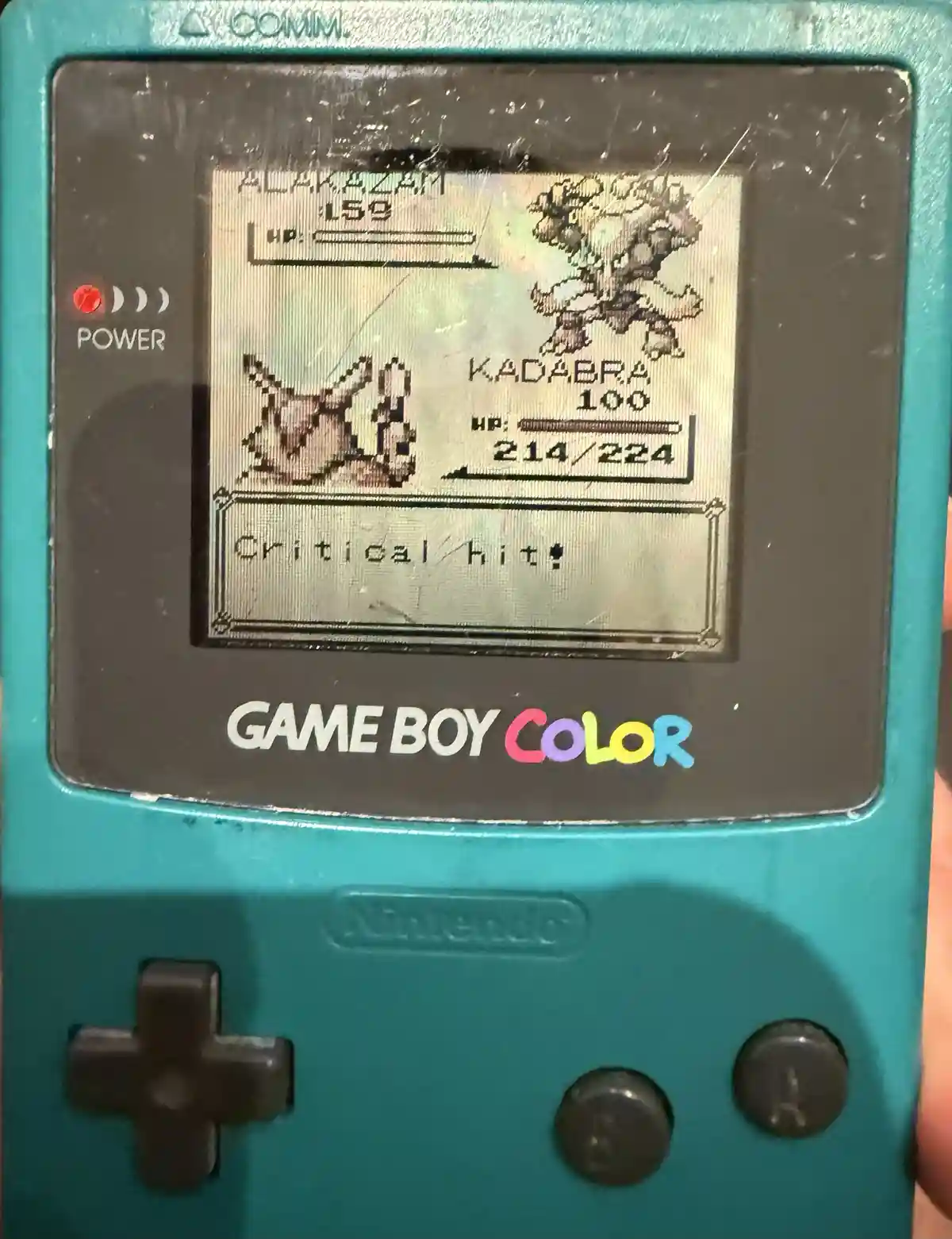 A teal Game Boy Color displays a Pokémon battle scene where Alakazam (level 59) attacks Kadabra (level 100), and the screen…