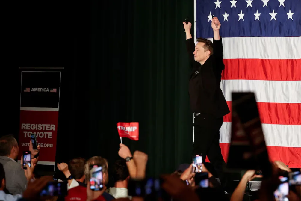 A man in a black shirt stands on a stage with arms raised, holding a microphone, in front of a large American flag. The…