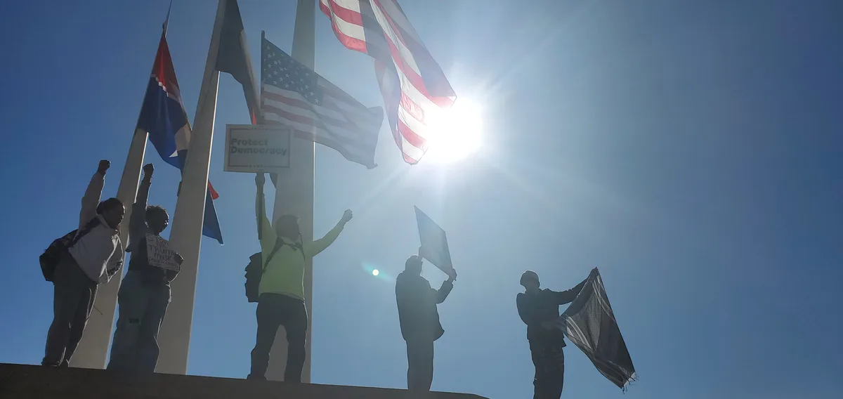 A low-angle photo shows silhouetted protesters holding signs and flags against a bright sun, with one sign reading 'Protect…