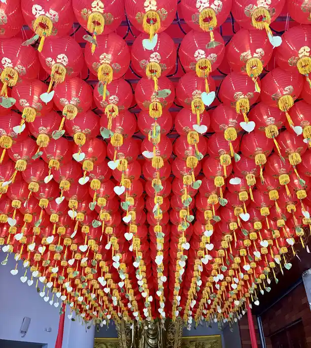 A dense ceiling of red Chinese lanterns with gold tassels and heart-shaped paper tags hanging down. The overlaid text reads…