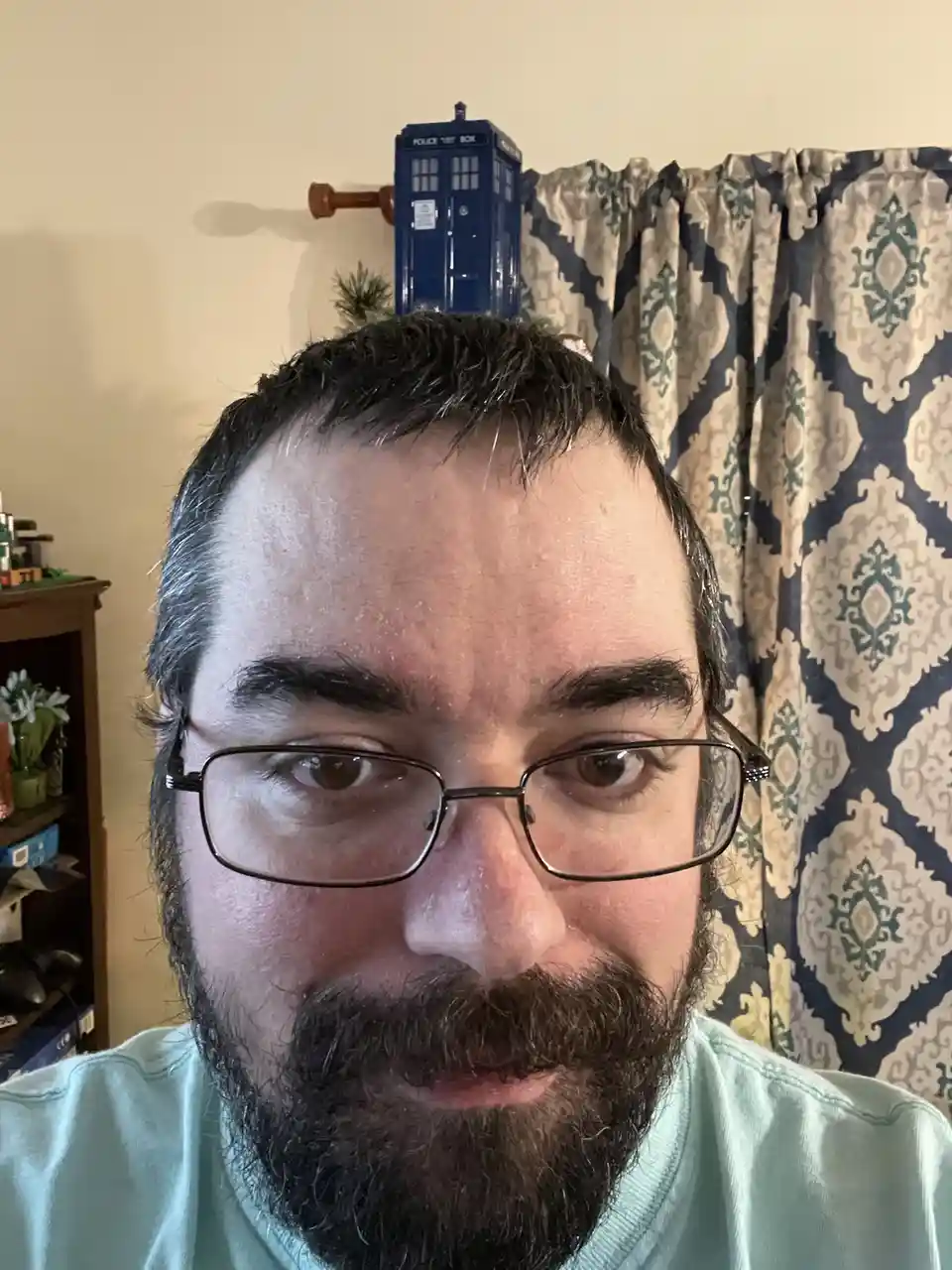 A man with glasses and a beard stares directly at the camera, with a small blue police box model (TARDIS) perched on top of…