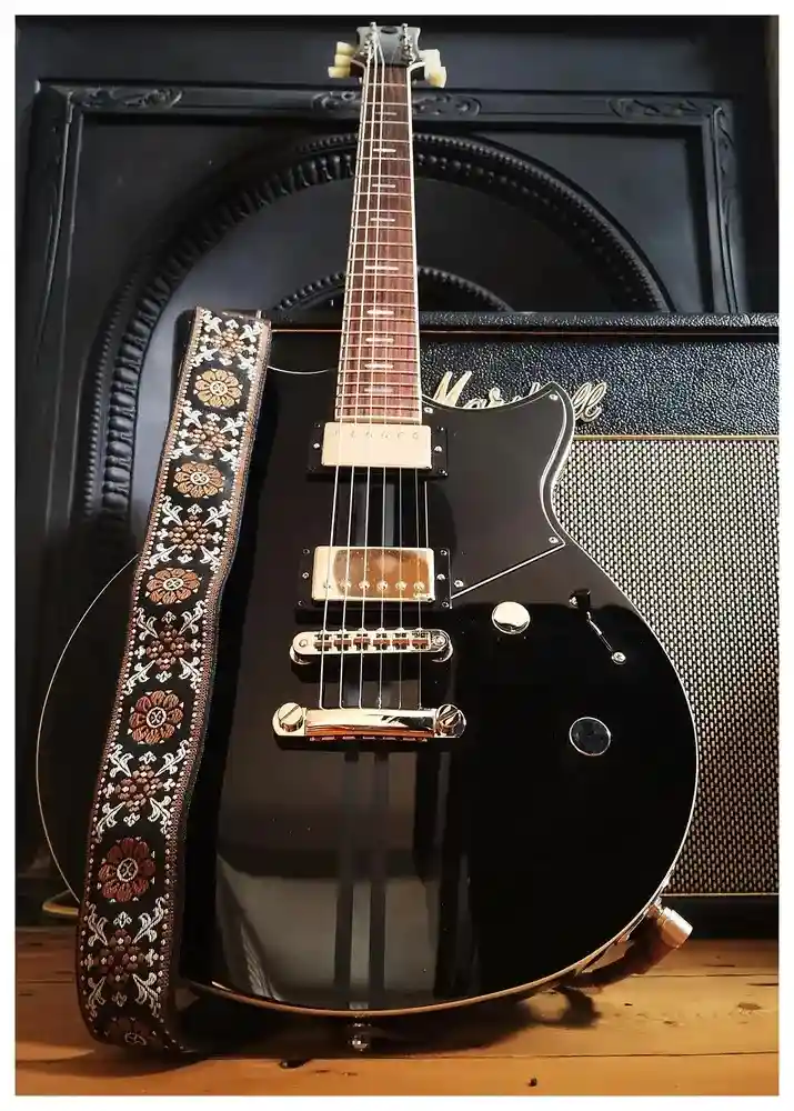 A black electric guitar with a decorative floral-patterned strap leans against a Marshall amplifier. The guitar has gold…