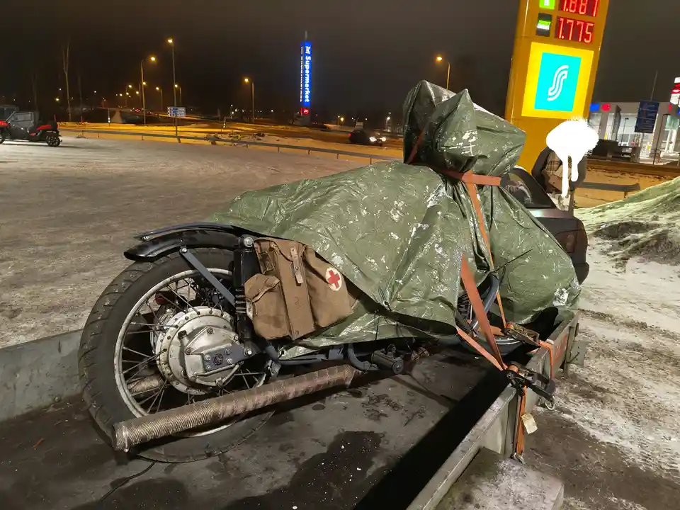 A motorcycle is fully covered with a green tarp and strapped down on a trailer at a snowy gas station at night. A brown…