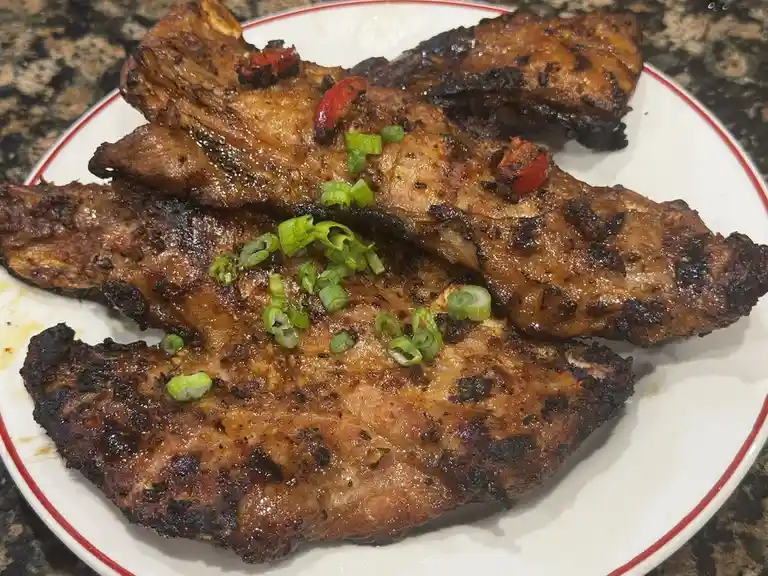A close-up photo shows several pieces of grilled meat on a white plate with a red rim, resting on a dark granite countertop…