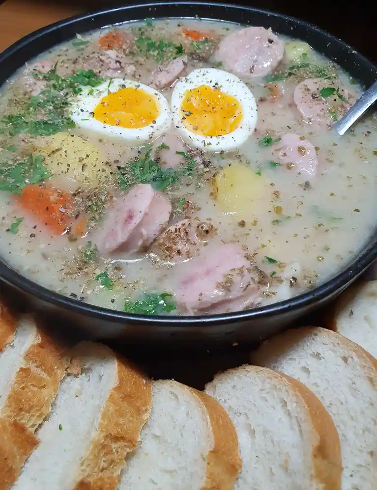A close-up photo shows a black bowl filled with creamy soup containing sliced sausages, chunks of potato, carrot pieces, and…