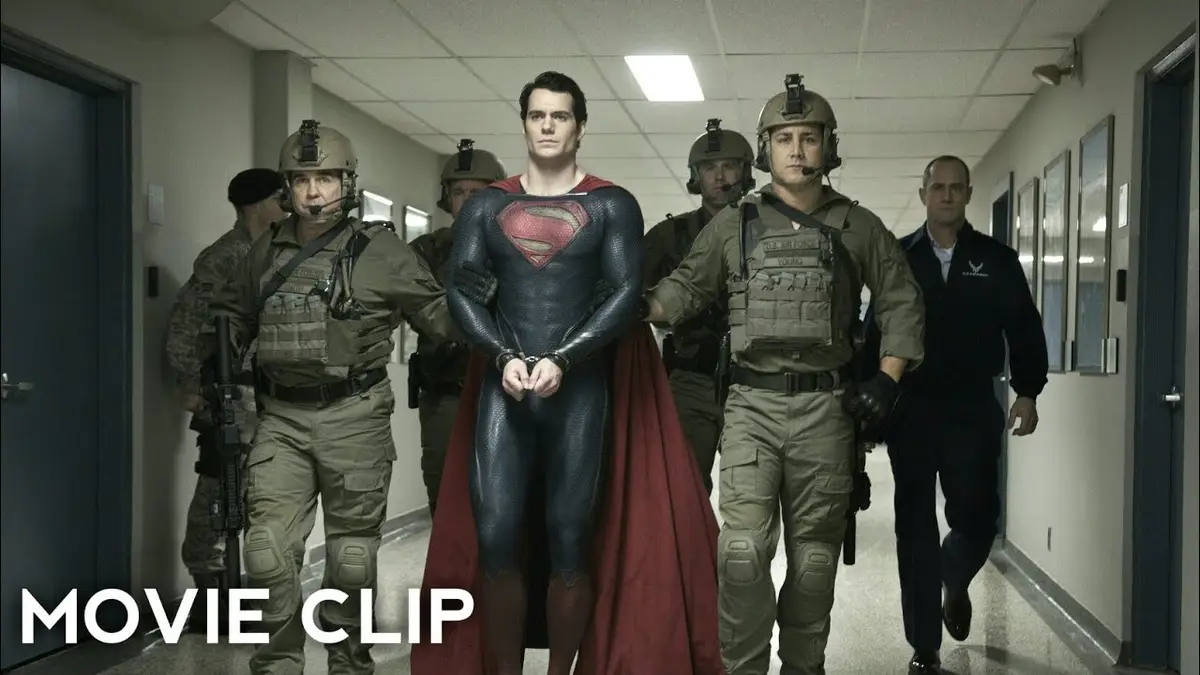 A still from a movie showing Superman, in his iconic red and blue suit with cape, being escorted down a sterile hallway by…