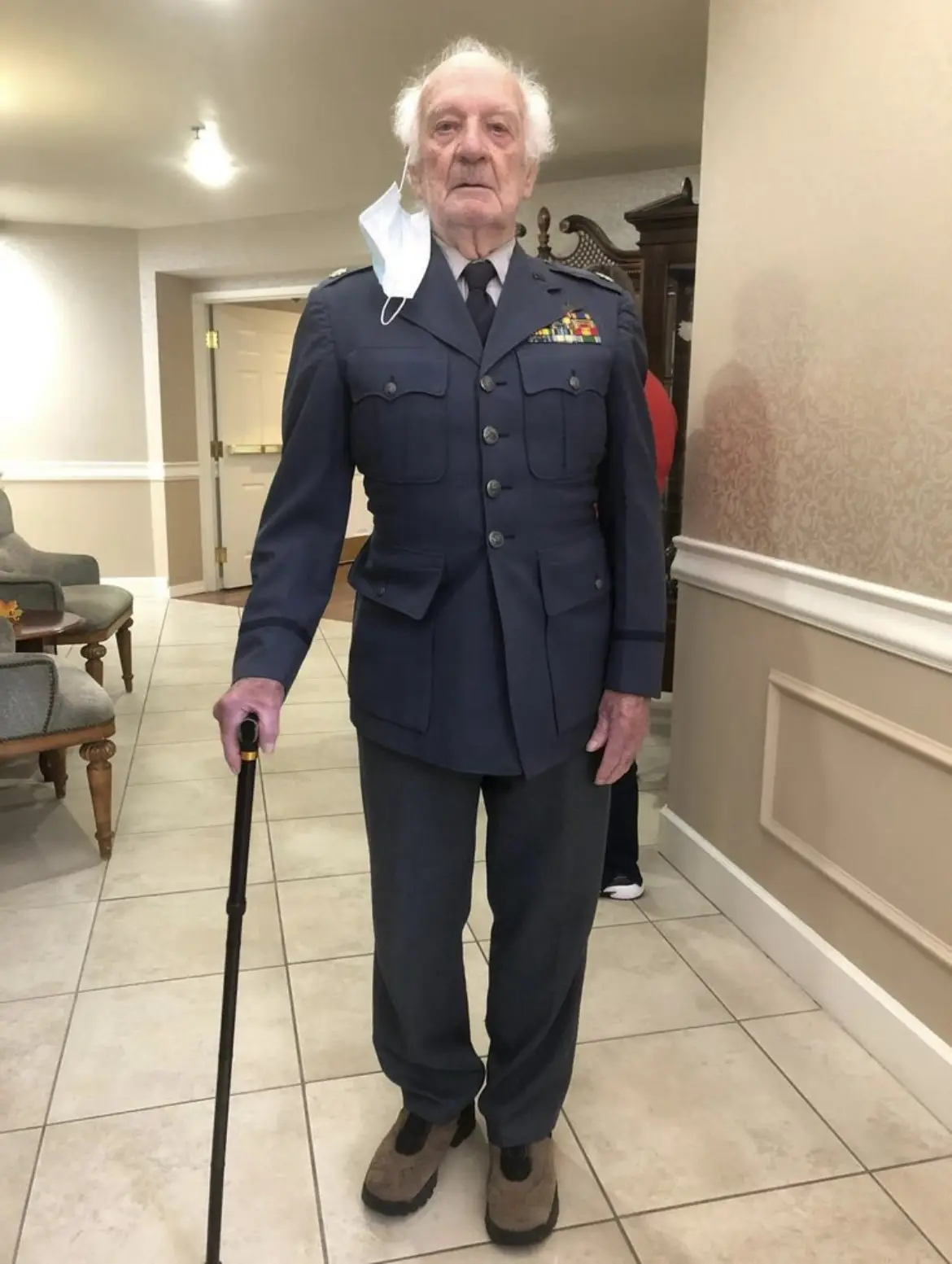 An elderly man in a formal military uniform with medals stands indoors holding a cane, a white face mask hangs from his…