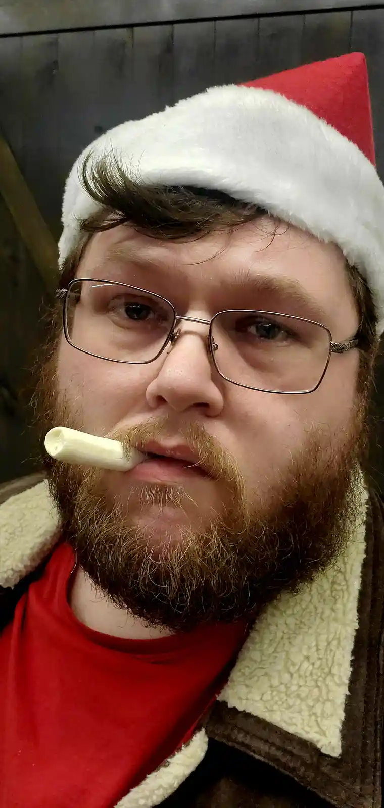 A man with glasses and a beard wears a red and white Santa hat and a brown jacket with a cream-colored fleece collar over a…