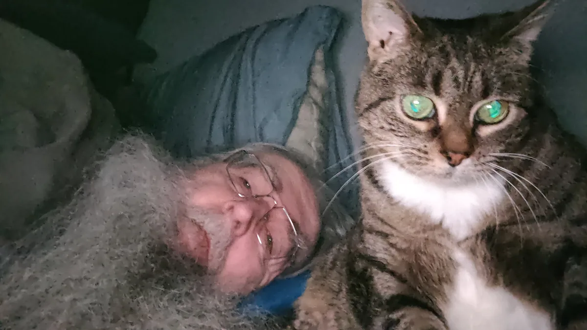 A man with glasses and a gray beard lies in bed next to a tabby cat with green eyes, both looking at the camera. The man is…