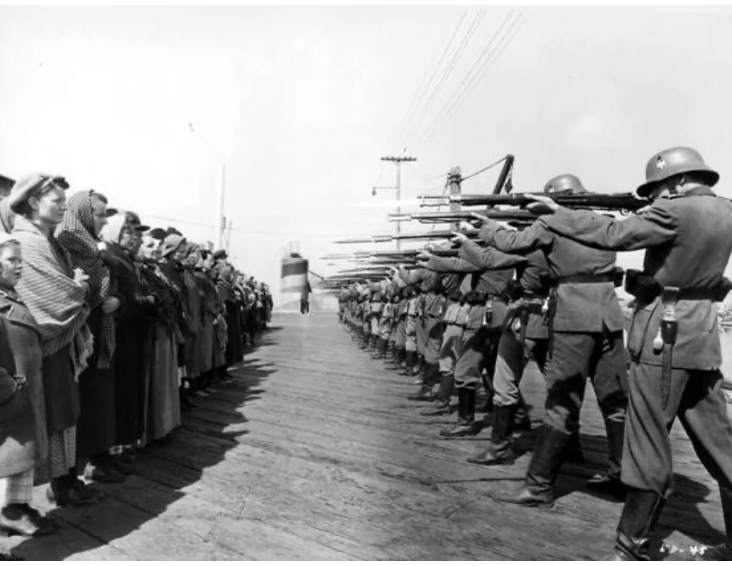 A black-and-white historical photo shows a line of soldiers in helmets and uniforms aiming rifles at a line of civilians…