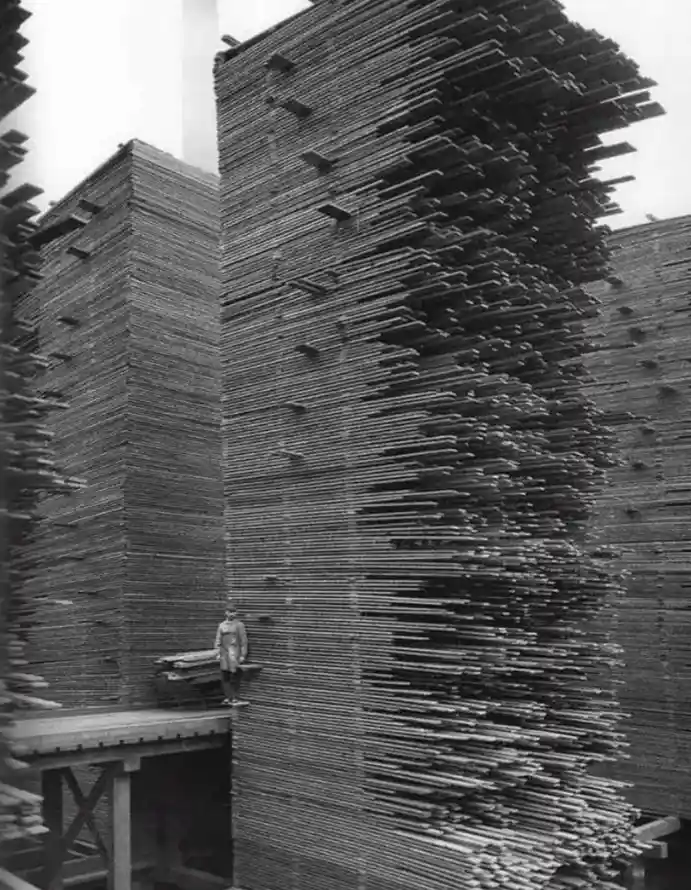 A black-and-white historical photo shows a person standing on a wooden platform next to towering stacks of lumber…