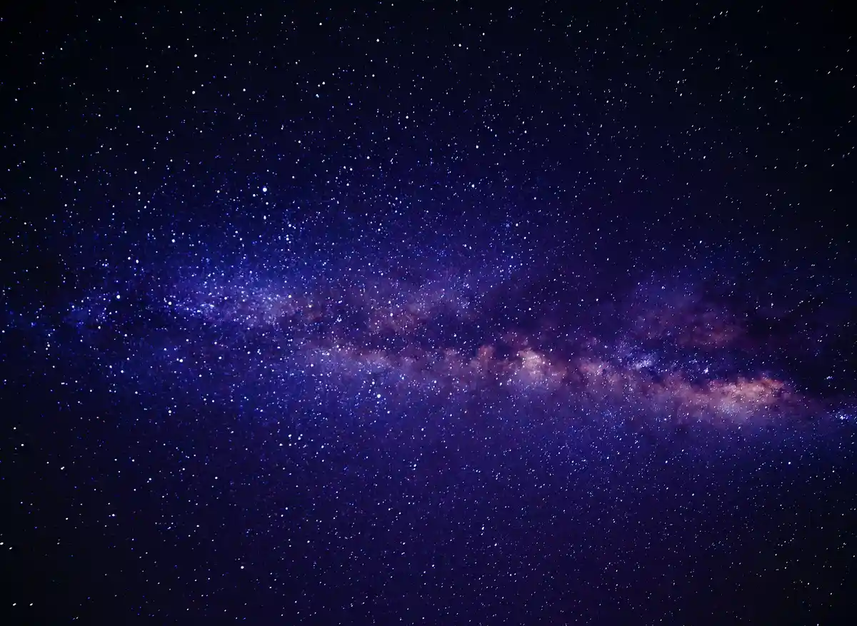 A stunning photograph of the Milky Way galaxy, showcasing a dense band of stars and cosmic dust in deep purple and blue hues…