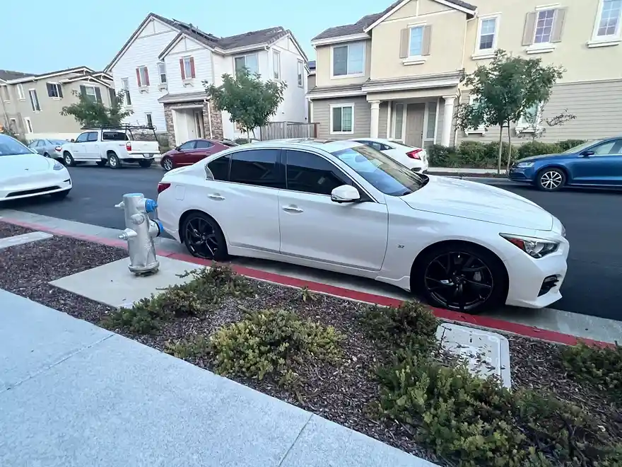 A white sedan with black rims is parked directly next to a silver fire hydrant on a residential street. The car is angled…