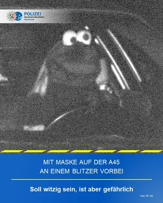 A grainy, black-and-white police dashcam photo shows a driver wearing a face mask while driving. The image has a blue banner…