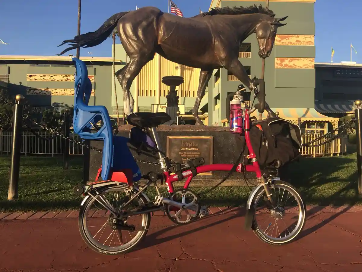 A red folding bicycle with a blue child seat is parked in front of a large bronze statue of a horse named Zenyatta, which…