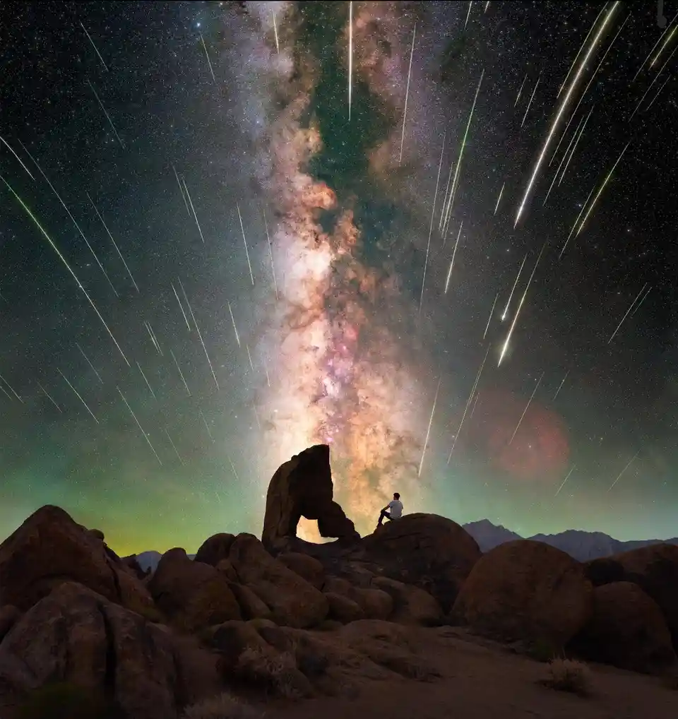 A person sits in silhouette atop large desert boulders, gazing up at a spectacular night sky filled with the Milky Way and…
