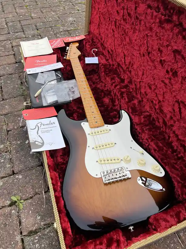 A Fender Stratocaster electric guitar with a sunburst finish sits inside its open, red-furred case on a brick patio. Beside…