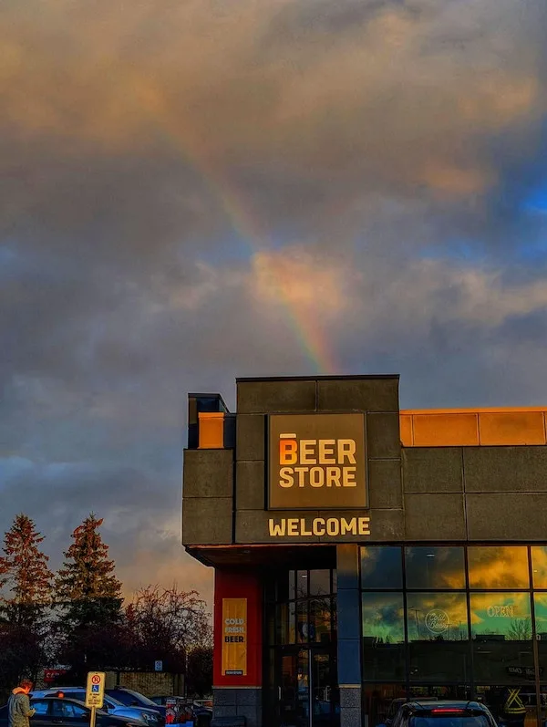 A photograph of a modern beer store building under a dramatic sky with a vivid rainbow arcing overhead. The store's sign…