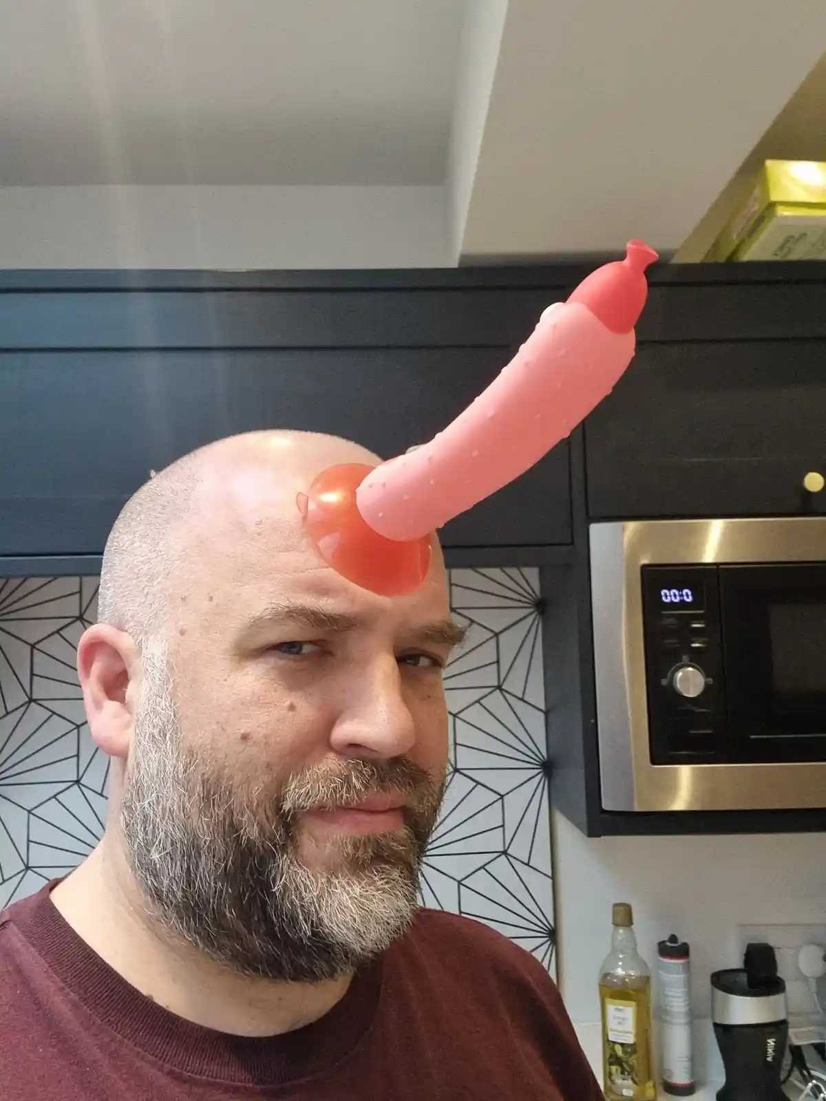 A bald man with a gray beard in a kitchen looks at the camera with a neutral expression while a pink, silicone sex toy is…