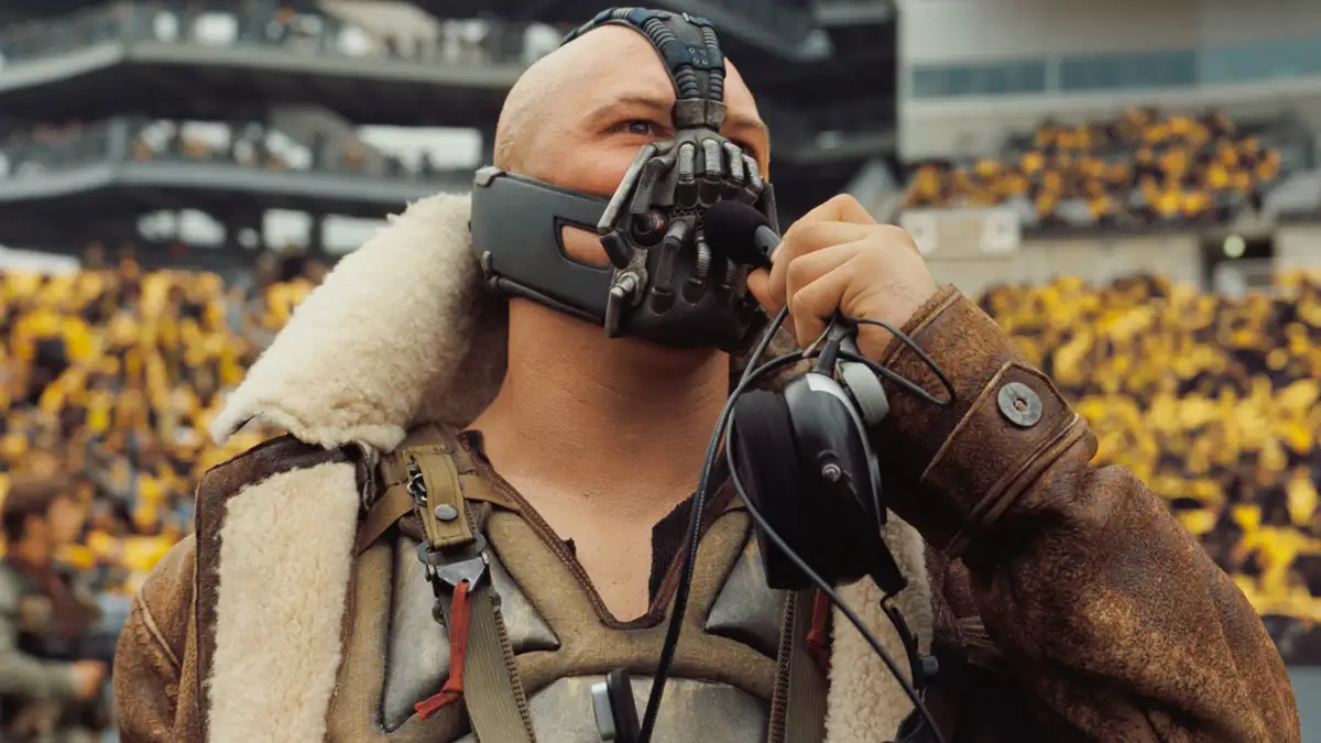 A bald man wearing a mechanical bat-like mask and a brown shearling-lined jacket holds a microphone and headset, standing…