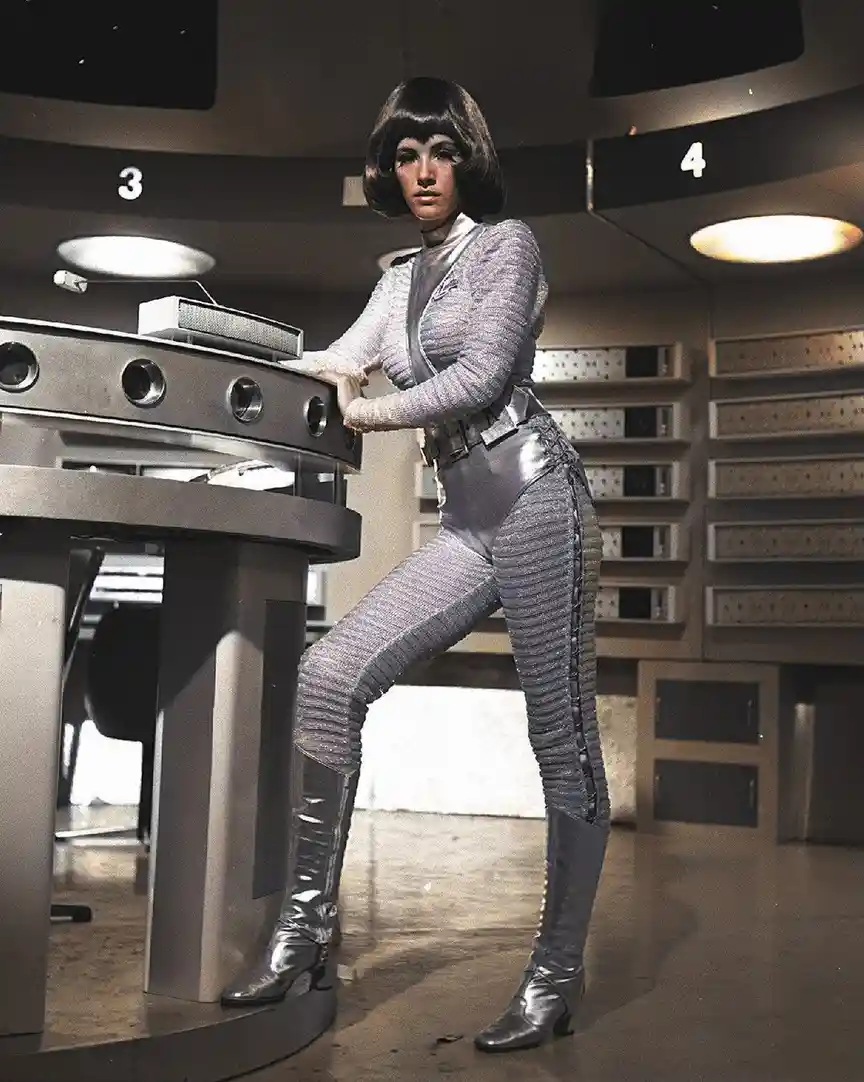 A woman in a silver, textured, form-fitting futuristic outfit with a bob haircut stands in a retro-futuristic control room…