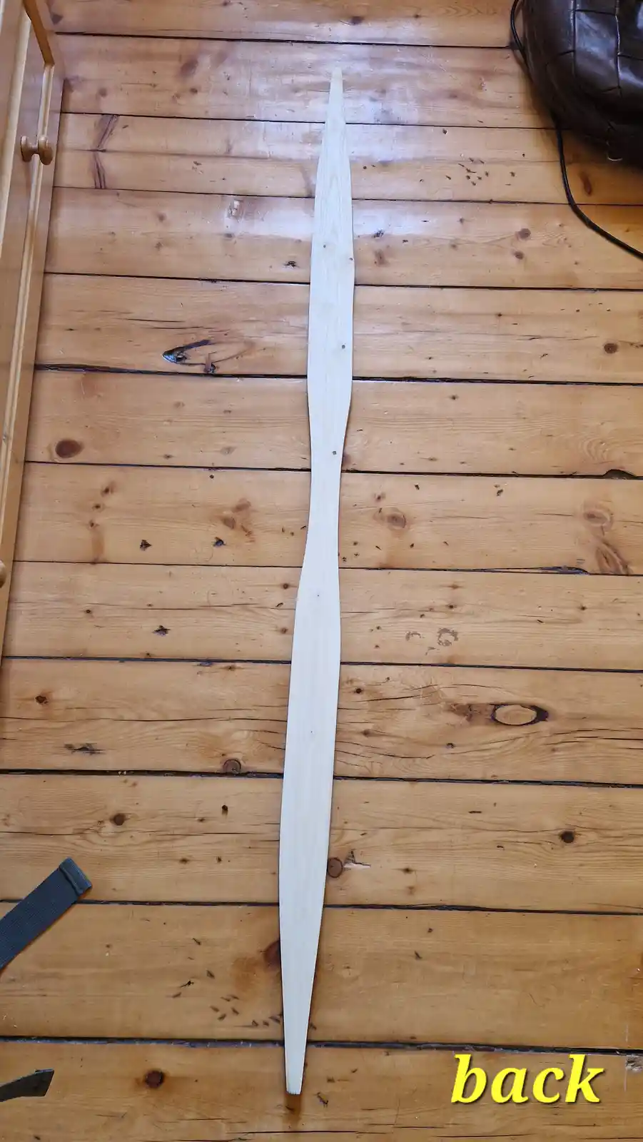 A long, light-colored wooden sword or blade lies vertically on a wooden floor, with the word 'back' in yellow text at the…