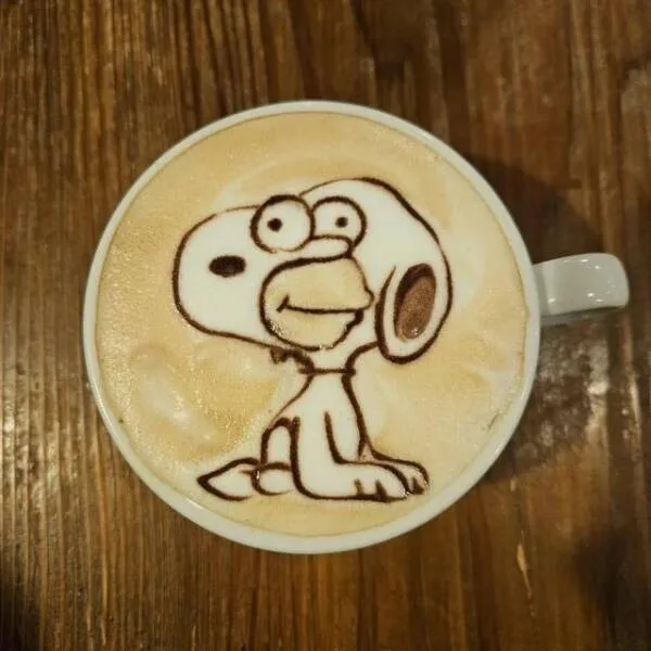 A top-down view of a white ceramic coffee mug on a wooden table, filled with a latte whose frothy surface has a detailed…