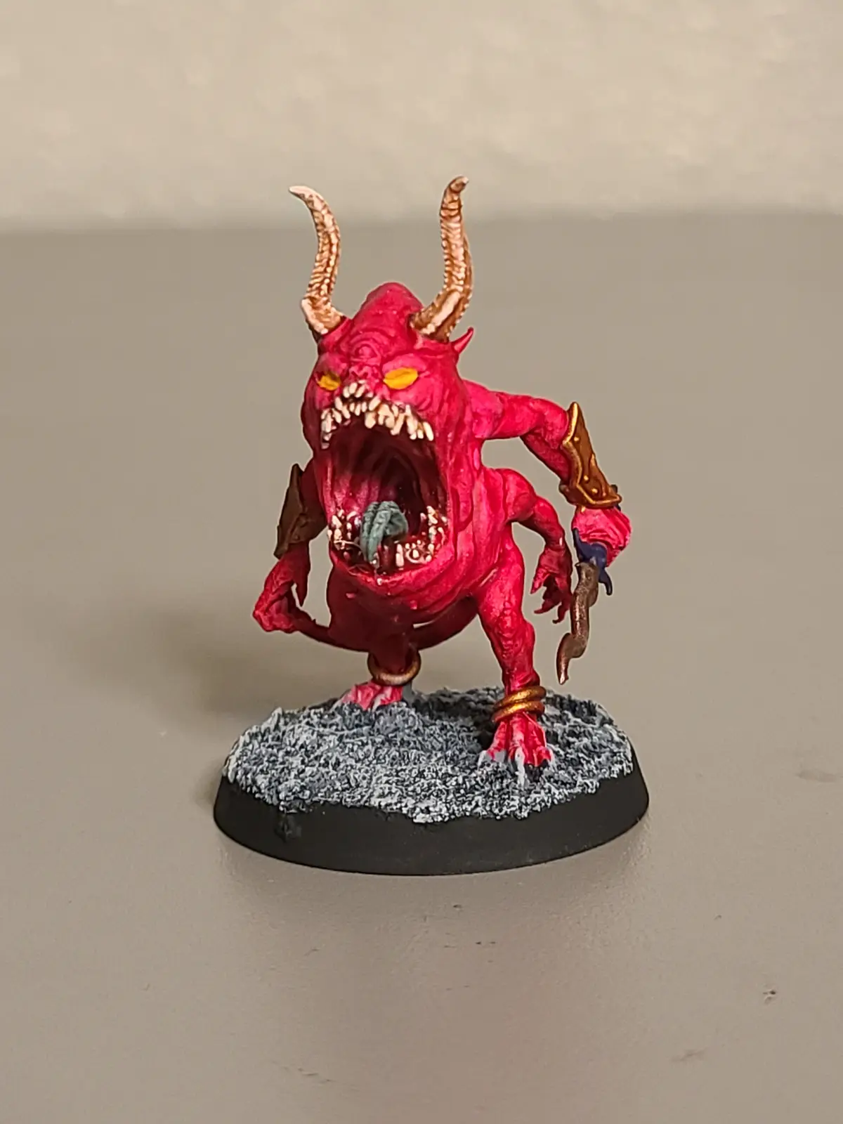 A hand-painted miniature of a red, horned demon with a wide-open mouth, sharp teeth, and glowing yellow eyes, standing on a…