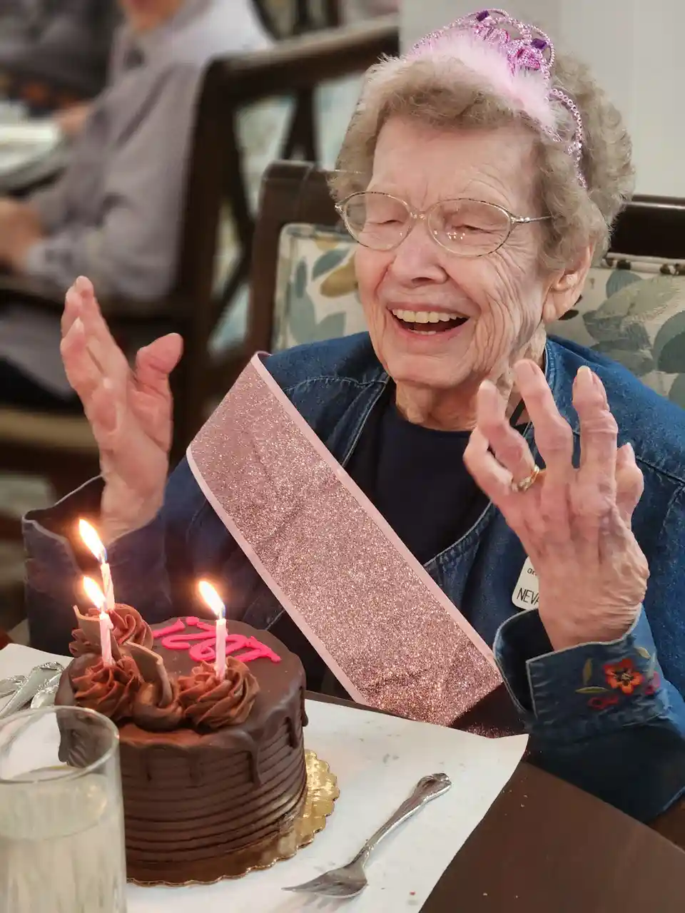 An elderly woman with glasses, wearing a pink glitter sash and a pink tiara, laughs joyfully with her hands raised in front…